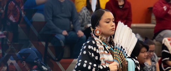 Young adult Native American female in traditional ceremonial dress standing in front of an audience.